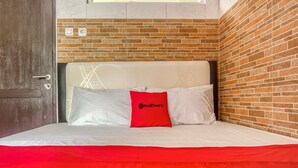 A hotel room with a brick-patterned wall, a bed with white bedding and a red accent pillow, and a door with a handle. - RedDoorz @ Panglima Polim 2 (Jakarta)
