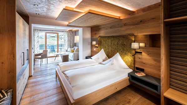 A modern hotel room with a wooden floor, a bed with white linens, a nightstand, a lamp, and a view of the outdoors.