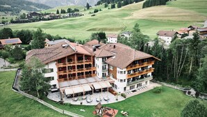 A large, multi-story building with a traditional Alpine architectural style, surrounded by lush green fields and mountains in the background.