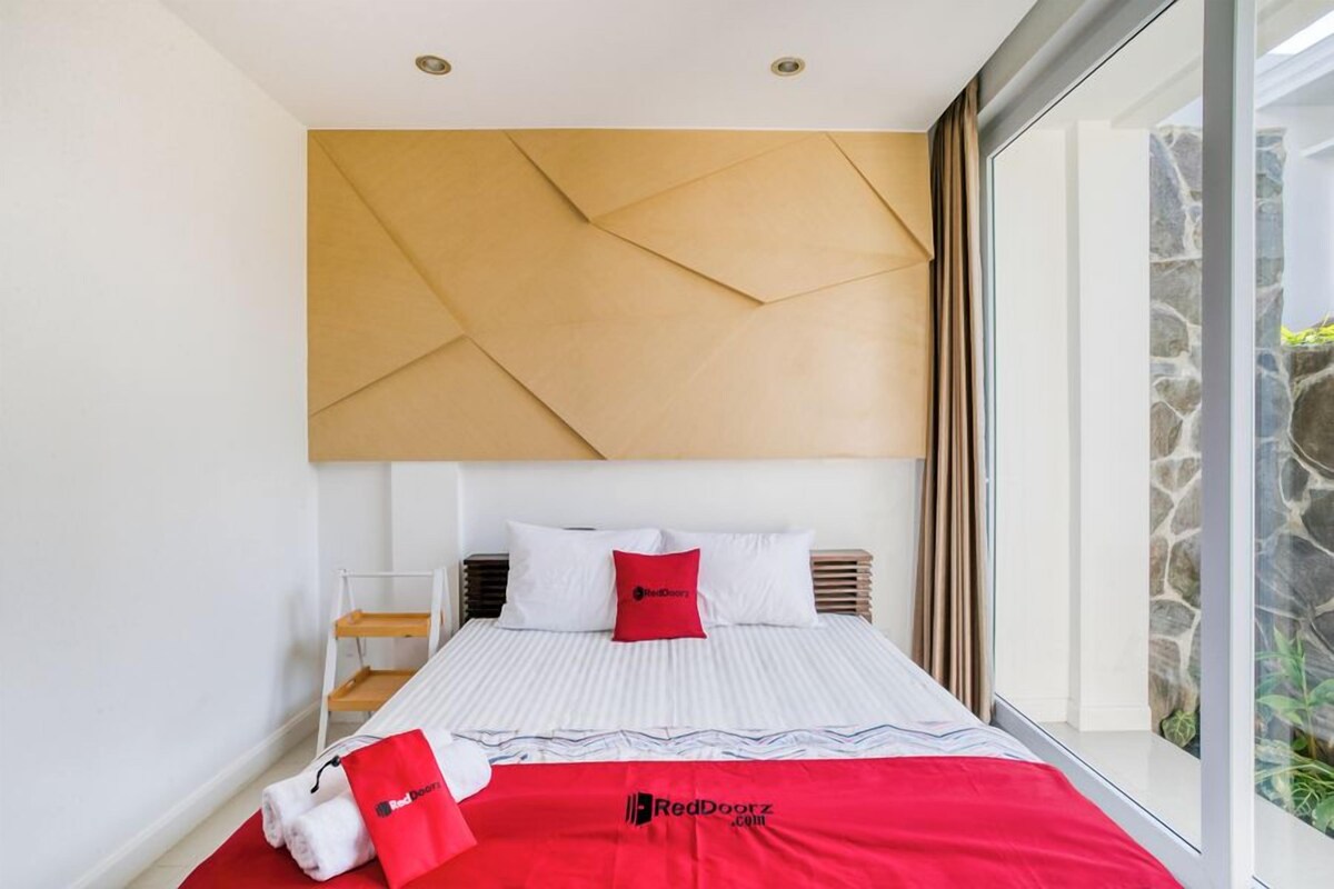 A hotel room with a large bed, a headboard, a window with a view of greenery, and a RedDoorz.com logo on the bedspread.