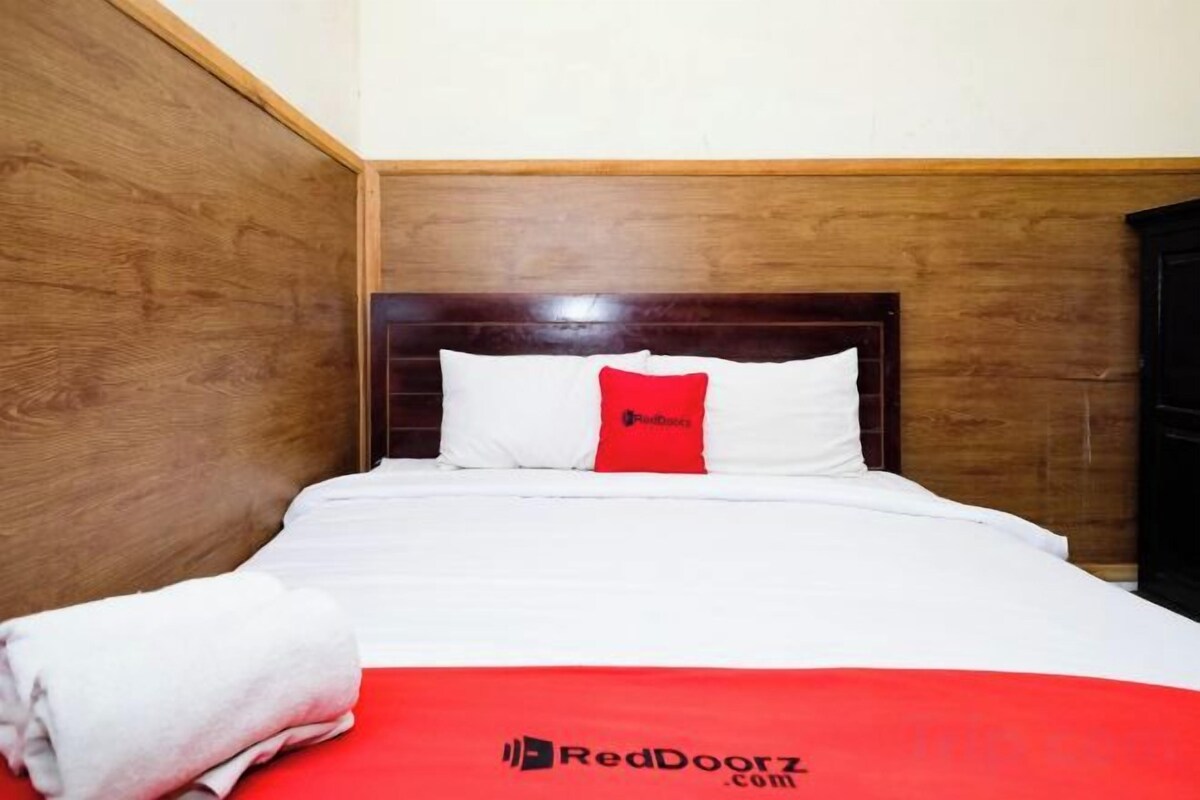 A hotel room with a wooden headboard, a white bed with a red pillow, and a red blanket with a logo.