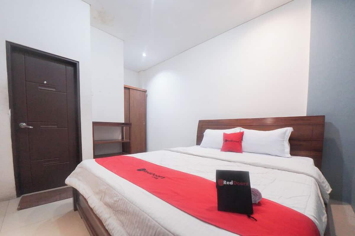 A hotel room with a bed, a wooden wardrobe, a door, and a red bedspread.