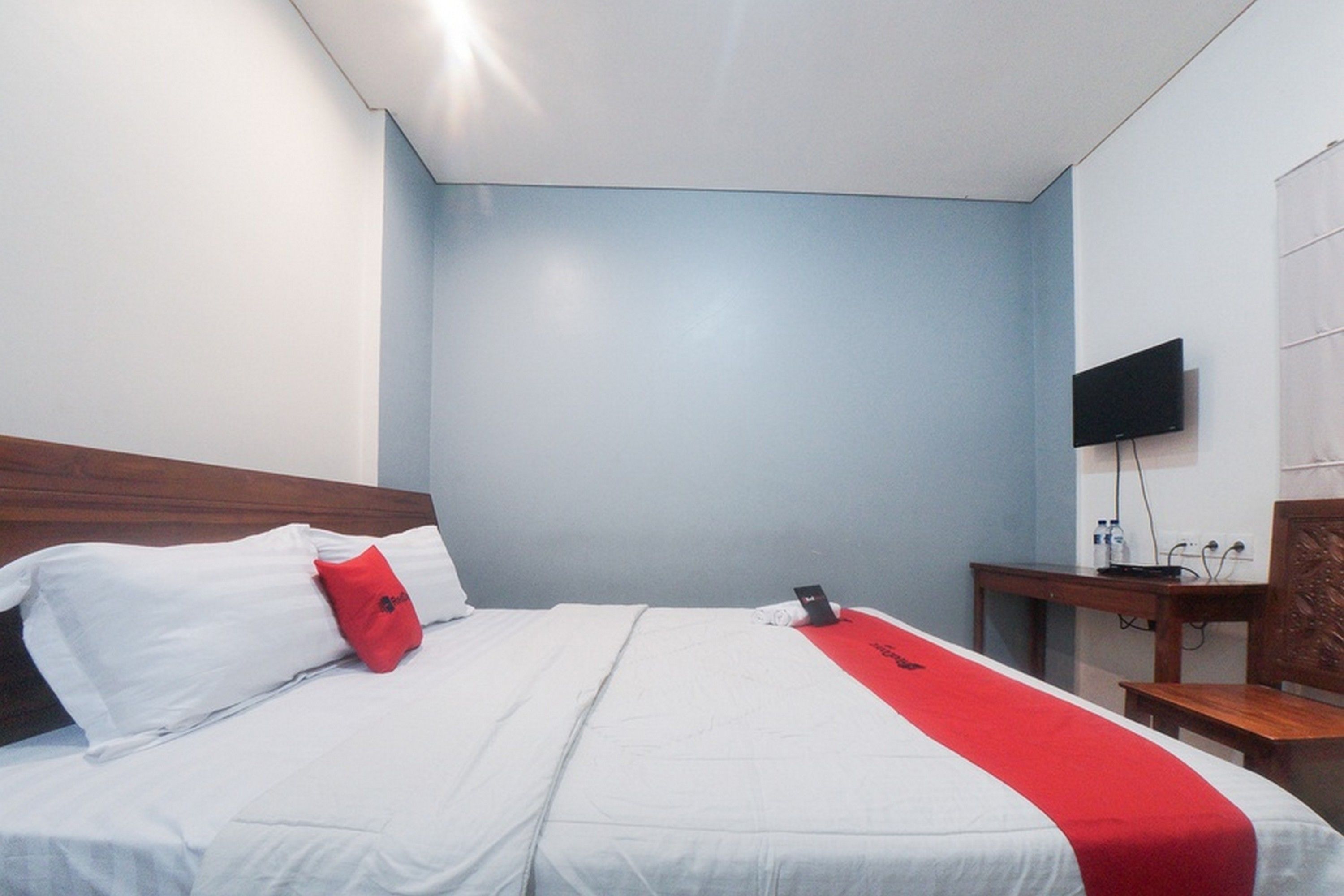 Habitación de hotel con cama, almohada roja, cabecero de madera, televisor montado en la pared y escritorio con silla.