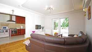 Una sala moderna con un sofá de cuero marrón, un televisor de pantalla plana y un comedor con armarios de madera, con una cocina al fondo.