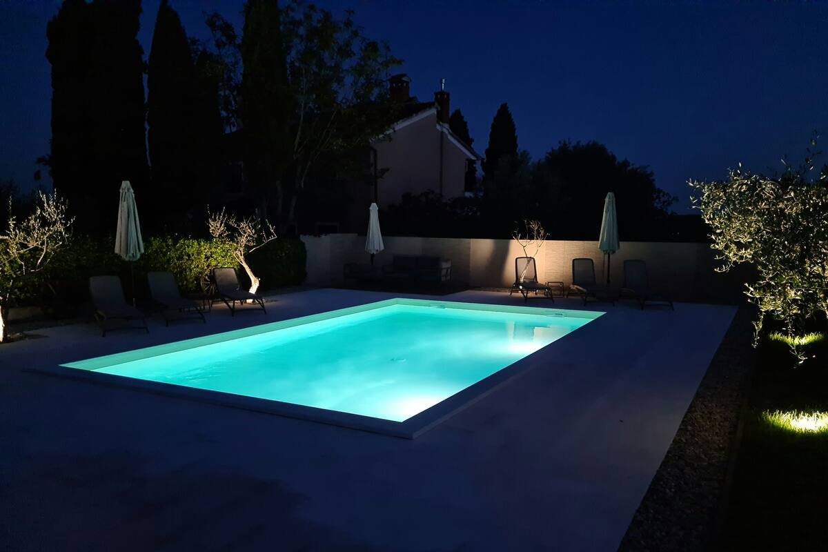 A rectangular swimming pool with a light blue tint, surrounded by a low wall. There are two lounge chairs and two umbrellas on either side of the pool.