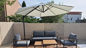 Um pátio ao ar livre com um guarda-sol bege, um sofá, duas poltronas e uma mesa de madeira.