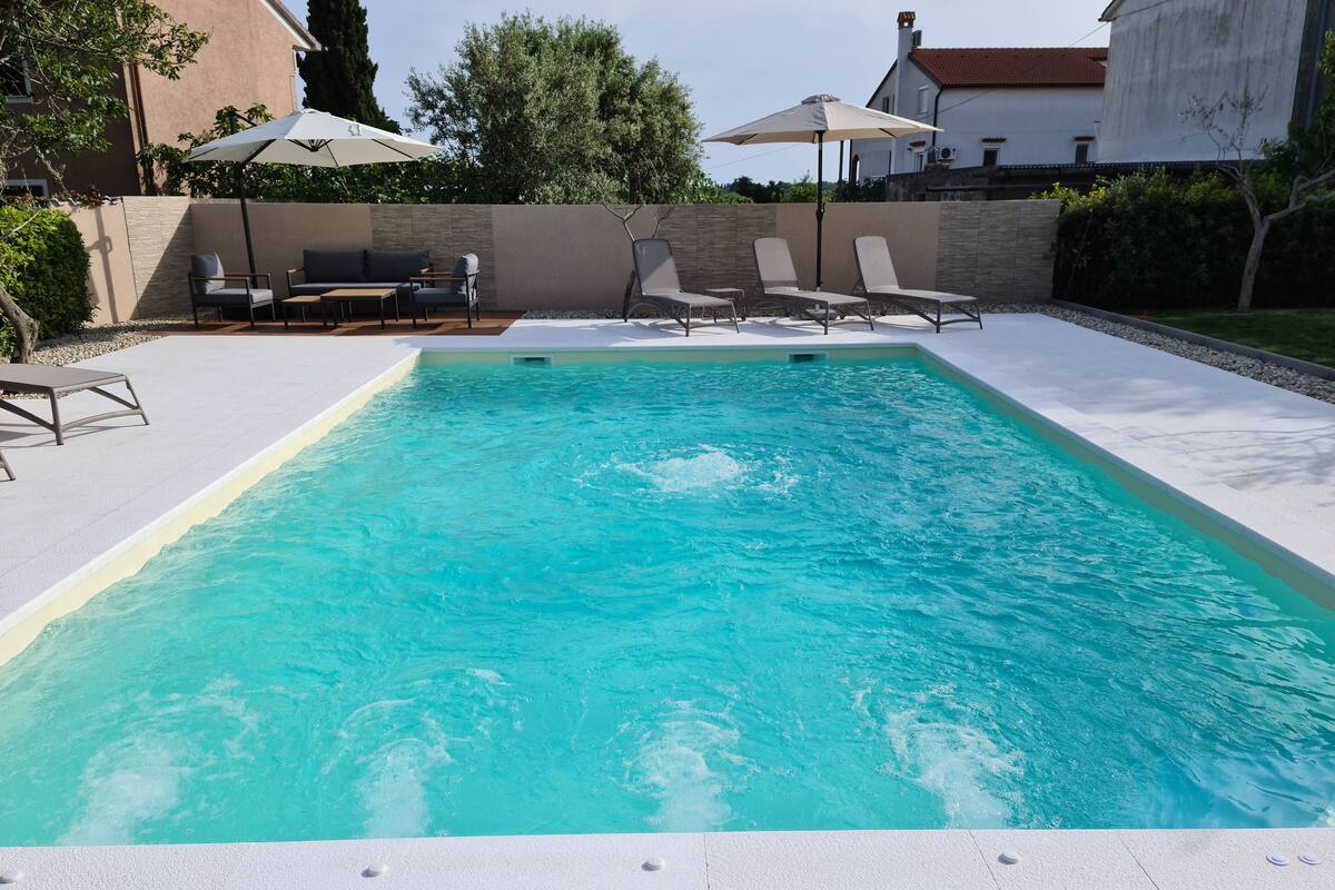 A rectangular swimming pool with clear blue water, surrounded by a white deck. There are lounge chairs and umbrellas in the background.