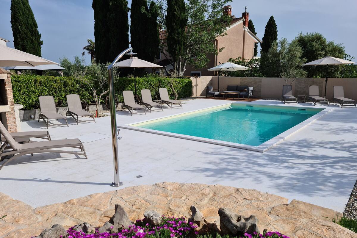 A swimming pool with lounge chairs and umbrellas, surrounded by stone walls and flowering plants.