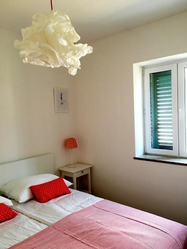 A bedroom with a bed, a nightstand, a red lamp, a white wall clock, and a window with blinds.