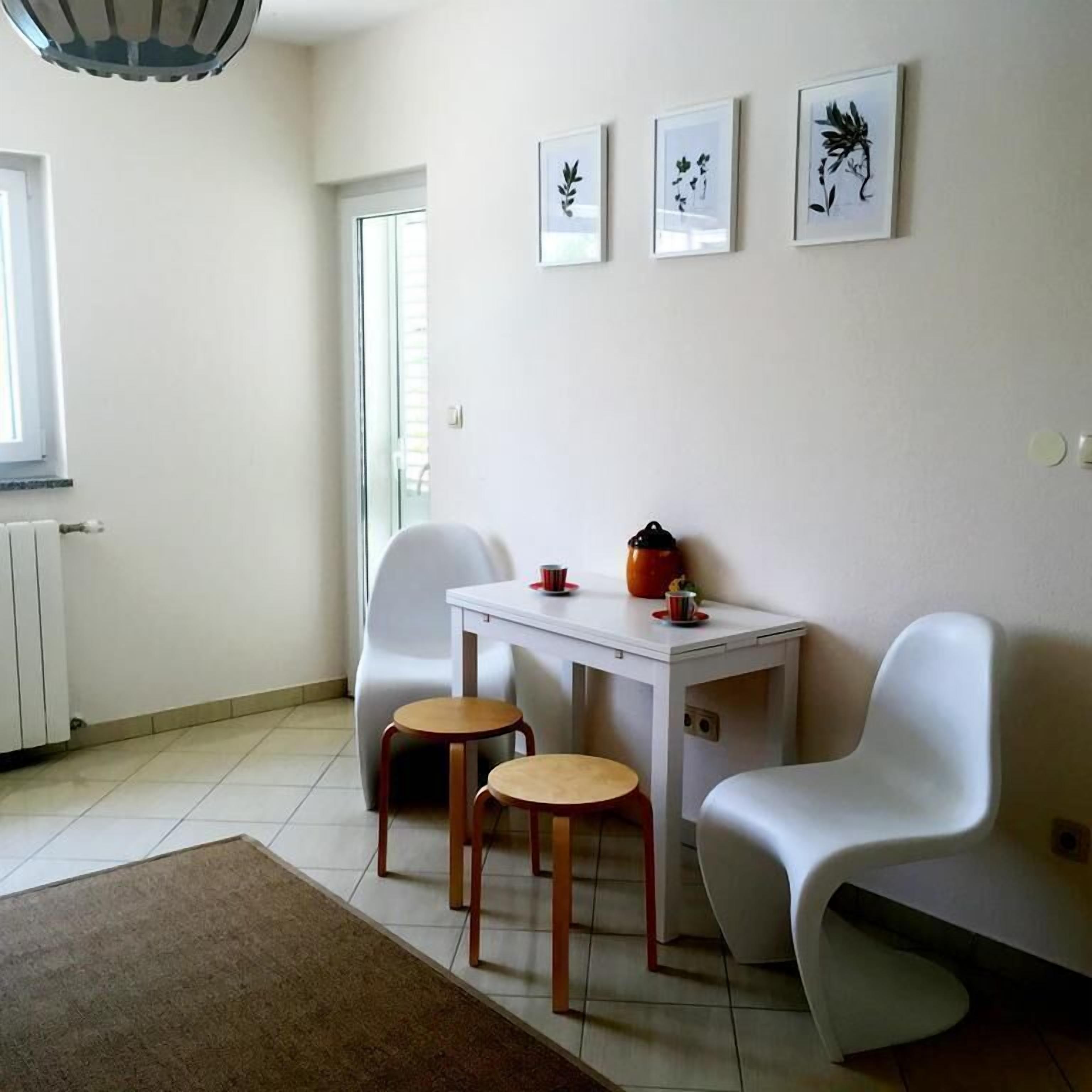 Ein kleiner, moderner Raum mit einem weißen Tisch, zwei Stühlen, einem Heizkörper und drei gerahmten Pflanzenbildern an der Wand.