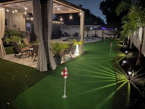 Un espacio exterior bien iluminado con césped artificial, piscina y una zona de estar cubierta con sillas y mesas.