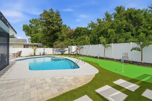 Una piscina con zona pavimentada alrededor, una hamaca y una sección de césped artificial verde.