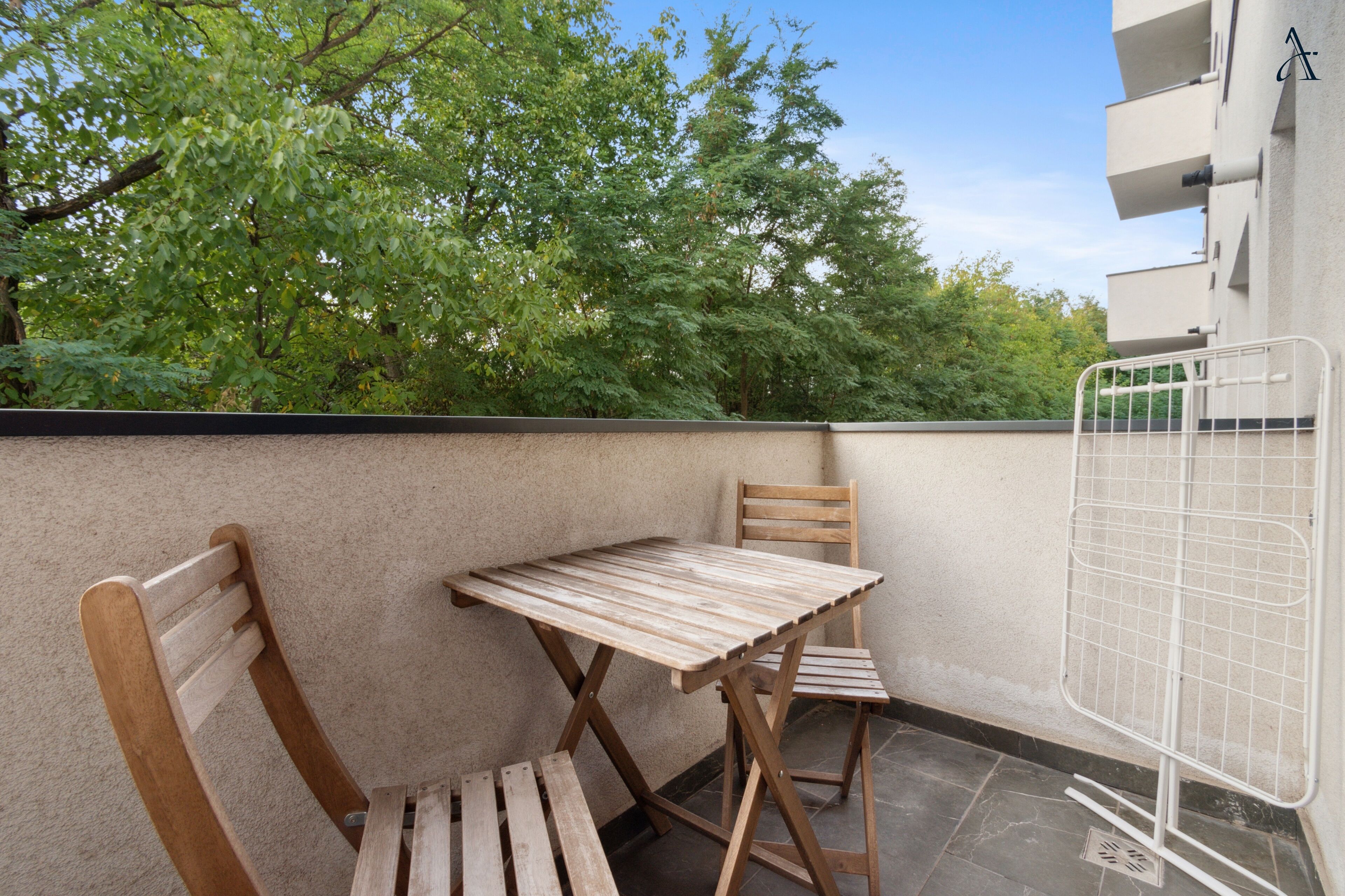 An outdoor balcony with a wooden table and two chairs, a metal gate, and a view of trees and buildings in the background.