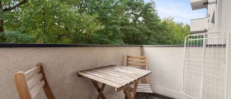 An outdoor balcony with a wooden table and two chairs, a metal gate, and a view of trees and buildings in the background.