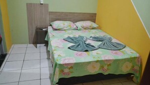 Quarto pequeno com cama de solteiro, colcha floral, cabeceira de madeira e mesa de cabeceira com controle remoto.