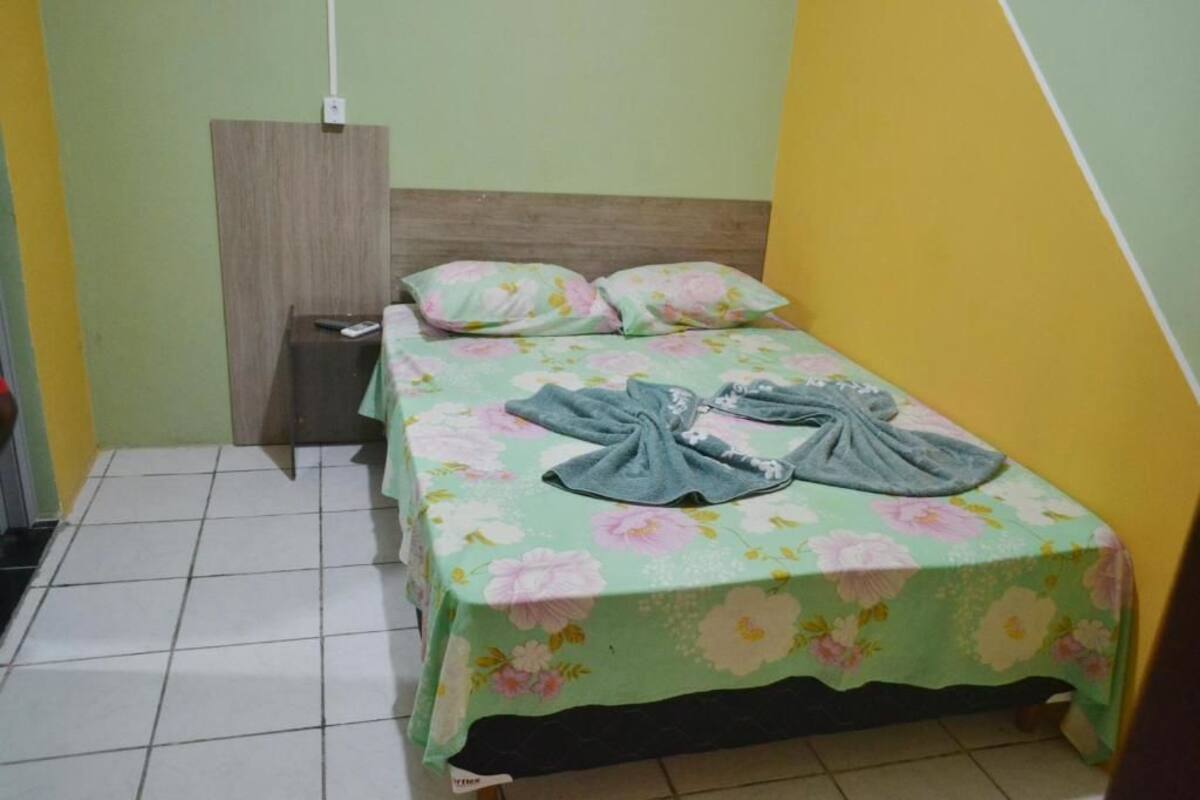 Ein kleines Zimmer mit einem Einzelbett, einer Bettdecke mit Blumenmuster, einem Holzkopfende und einem Nachttisch mit Fernbedienung.