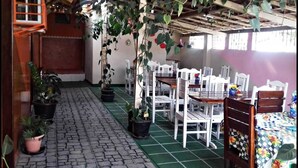 Uma área de refeições ao ar livre coberta, com mesas, cadeiras, plantas em vasos e piso de pedras irregulares.