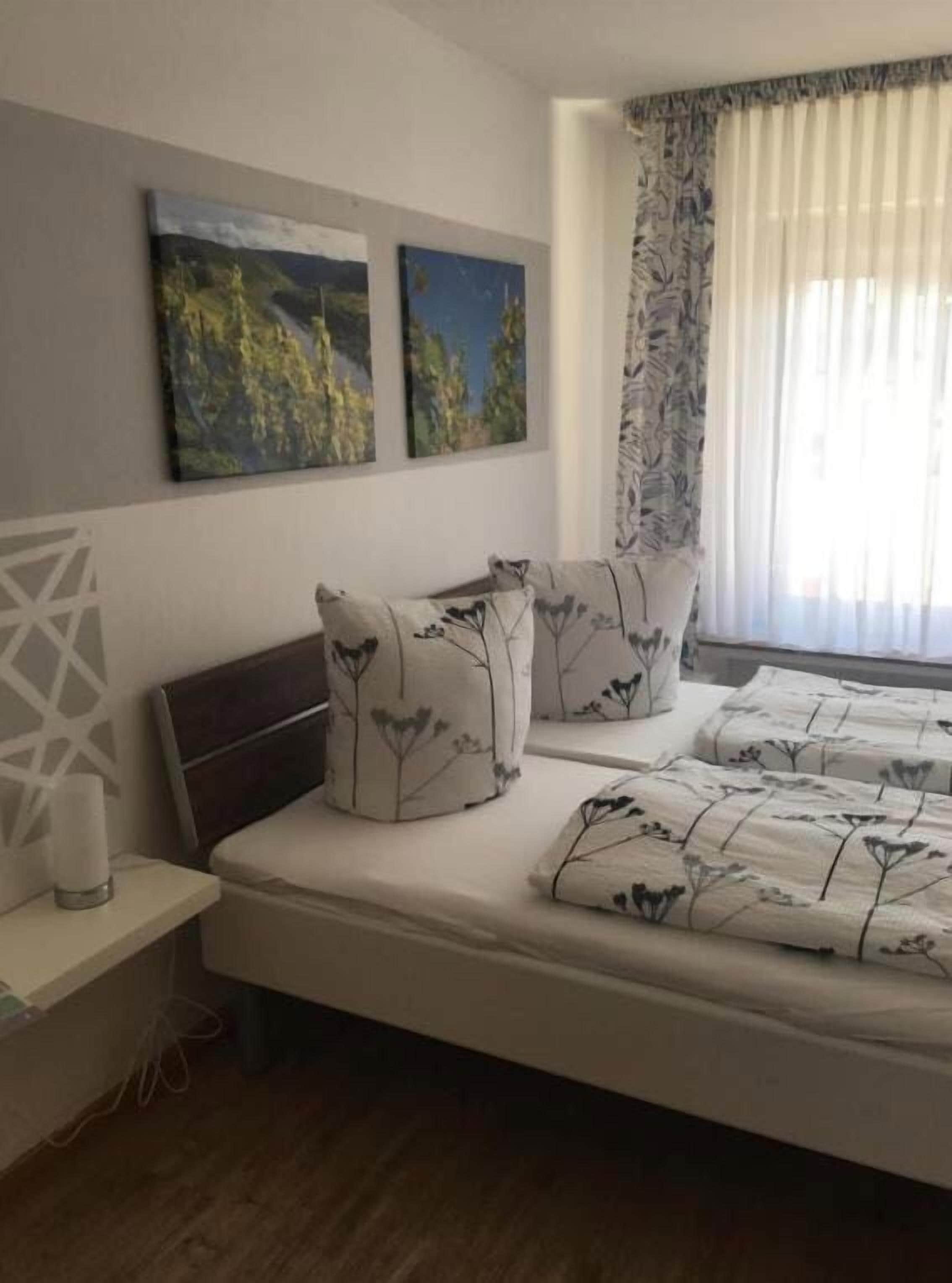 Une chambre à coucher avec un lit, deux tableaux accrochés au mur, une table de chevet avec une lampe et une fenêtre avec des rideaux.