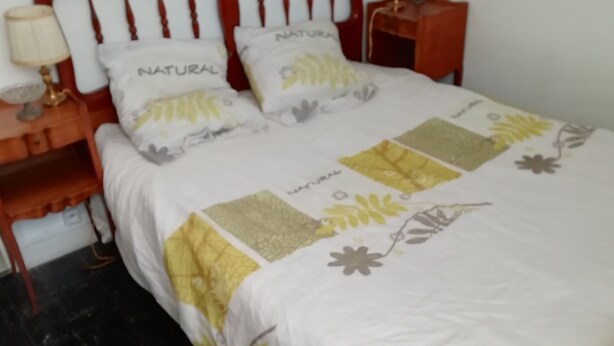 Ein Bett mit einer weiß-gelben blumigen Tagesdecke, zwei Kissen mit dem Schriftzug 'NATURAL', ein hölzernes Kopfteil und ein Nachttisch mit einer Lampe.