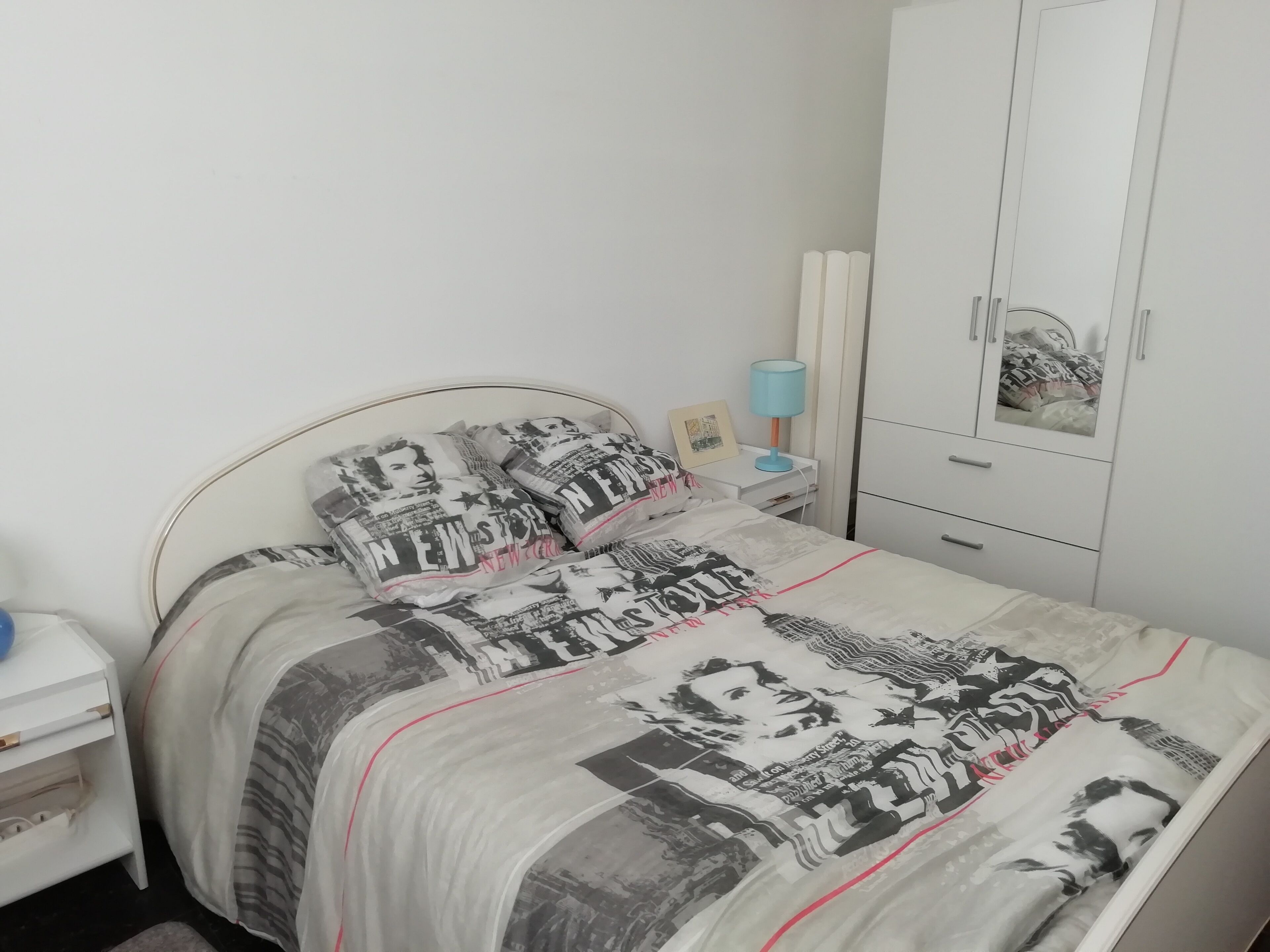 A bedroom with a bed featuring a patterned bedspread, a bedside table with a lamp, and a wardrobe.