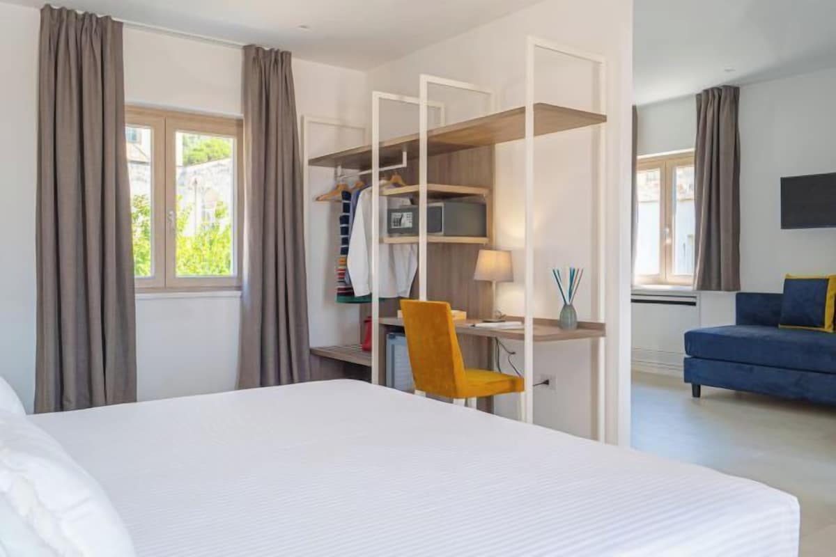 A hotel room with a bed, a desk with a chair, a wardrobe, and a sofa.