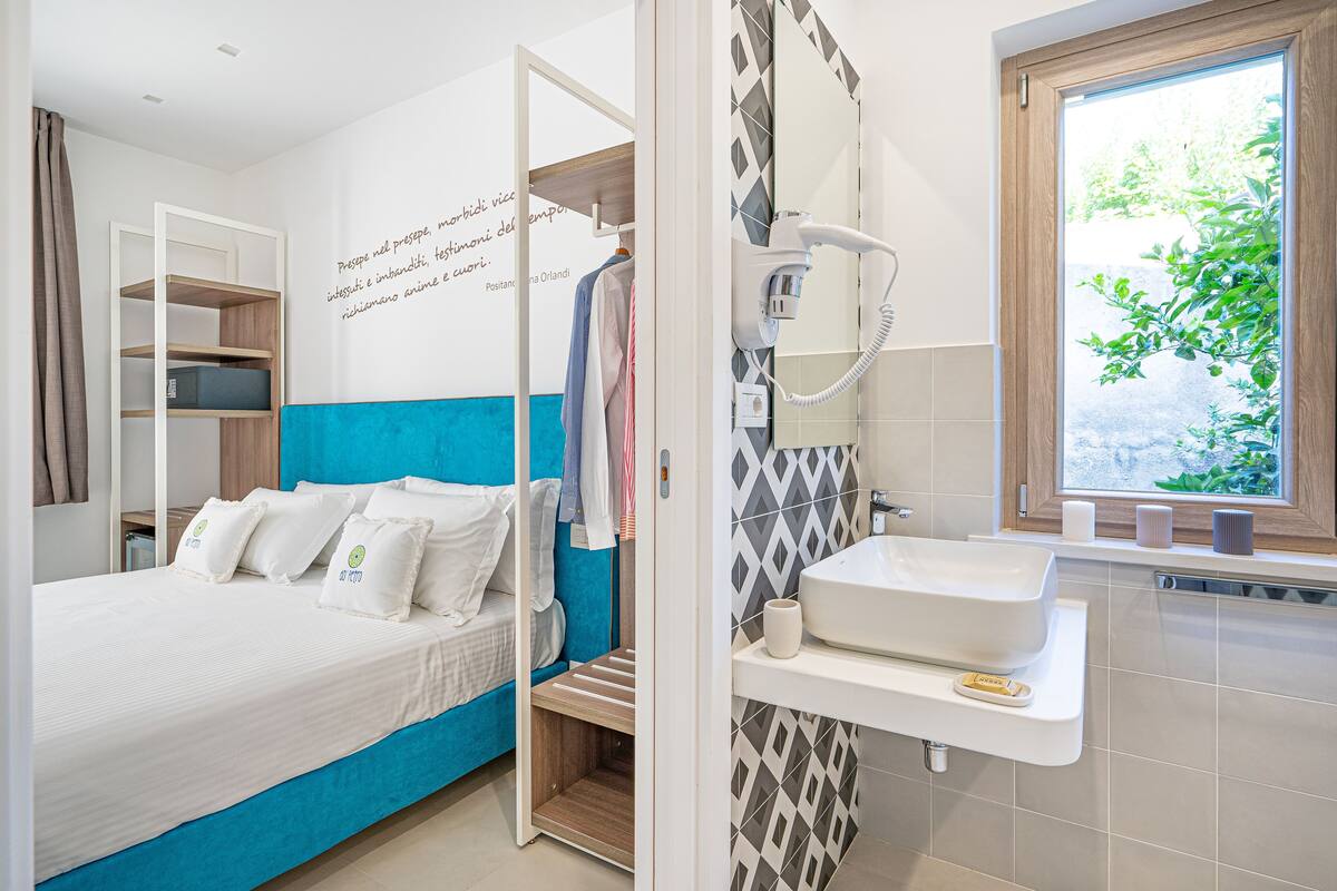 A modern hotel room with a bed, a built-in wardrobe, a bathroom with a sink and mirror, and a window with a view of greenery.