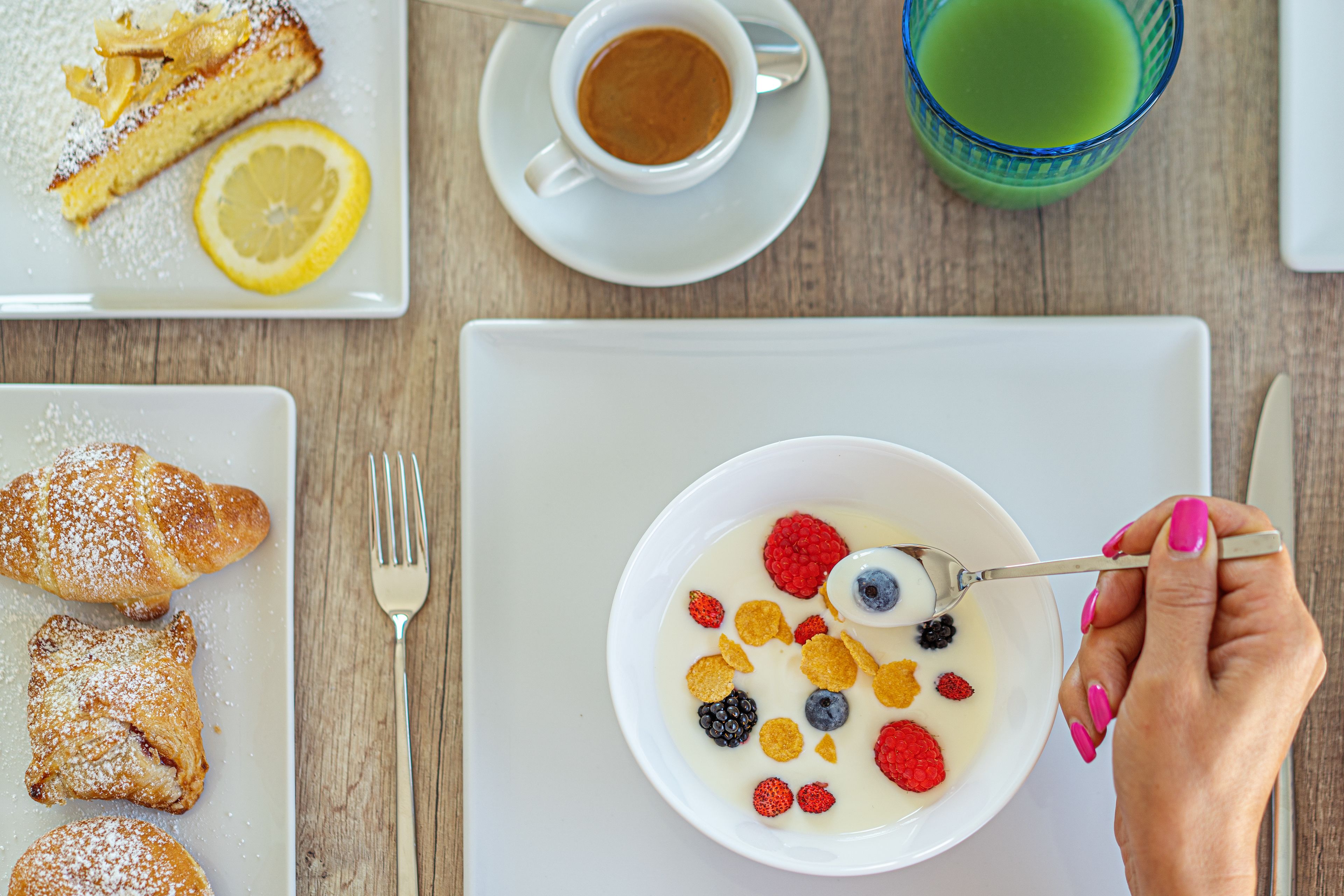 A breakfast setting with a plate of pastries, a bowl of yogurt with berries, a cup of coffee, and a glass of green juice.