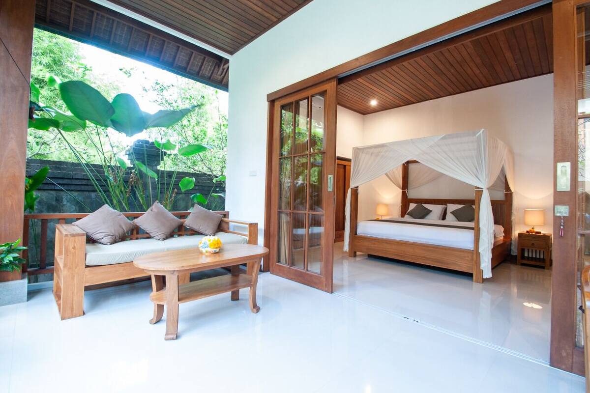 A room with a wooden sofa, a bed with a canopy, a small table, and a view of greenery outside.