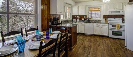 A kitchen with white cabinets, a wooden dining table set for four, and a large window.