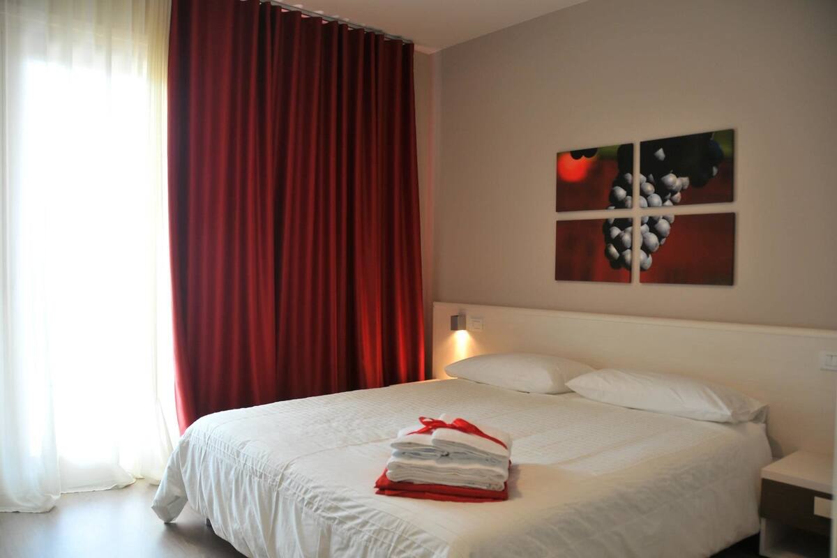 Un lit bien fait, avec du linge de lit blanc et une serviette rouge, une table de chevet et un mur orné d’une œuvre d’art représentant des raisins.
