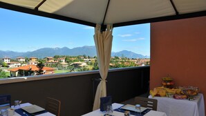Eine Terrasse mit gedeckten Tischen, Blick auf eine Stadt und Berge, sowie ein Tisch mit Speisen und Getränken.