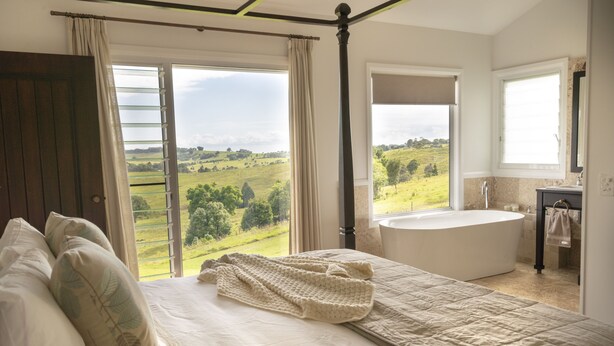 A bedroom with a large bed, a view of a green landscape, a bathtub, and a window with curtains. - Enjoy this piece of paradise and immerse yourself in luxury and style. (Byron Bay)
