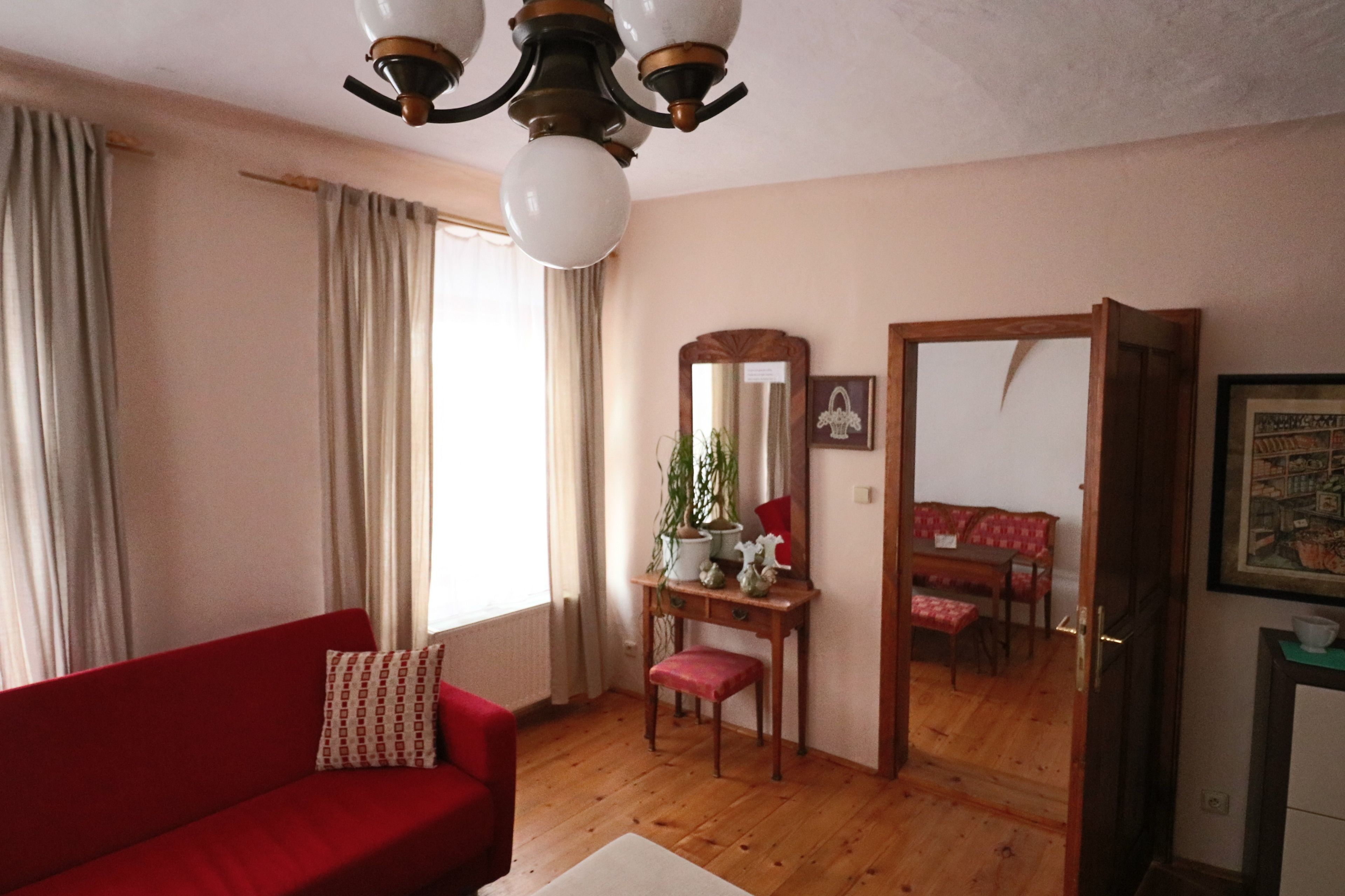 A room with a red sofa, a wooden desk with a mirror, a dining table with chairs, and a ceiling fan.