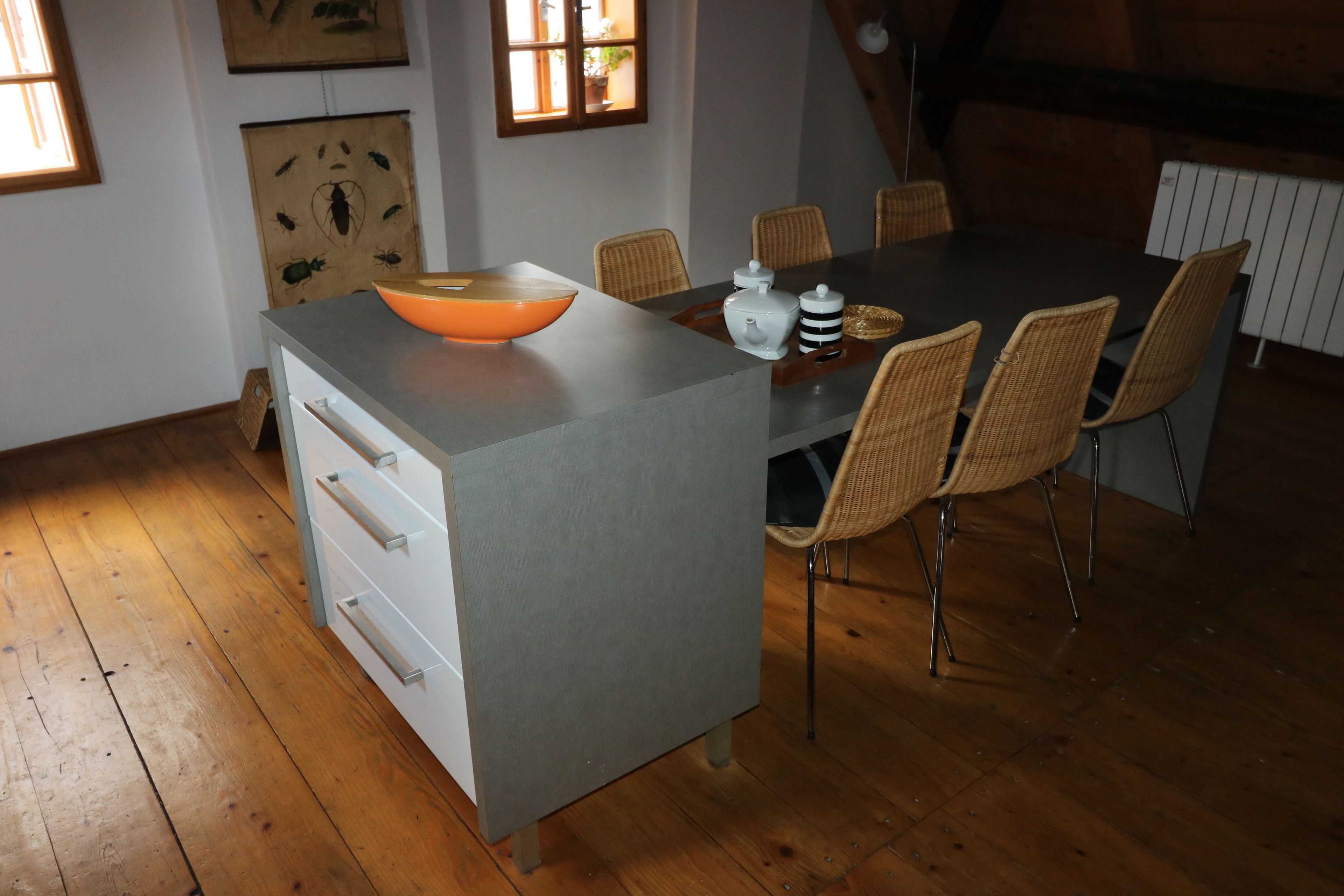 A dining area with a grey table, wicker chairs, and a white cabinet with drawers.