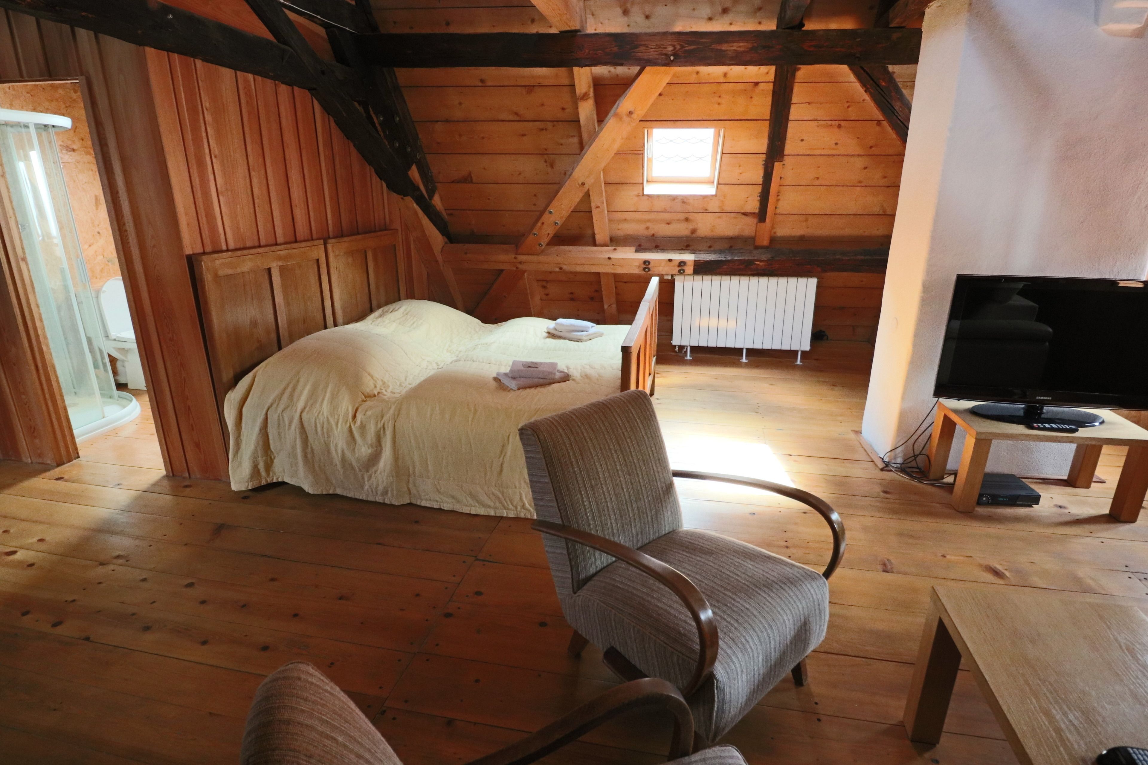 Ein gemütliches Schlafzimmer im Dachgeschoss mit Holzbalkendecke, einem Bett mit weißer Bettdecke, einem Holzstuhl, einem kleinen Tisch und einem Fernseher.