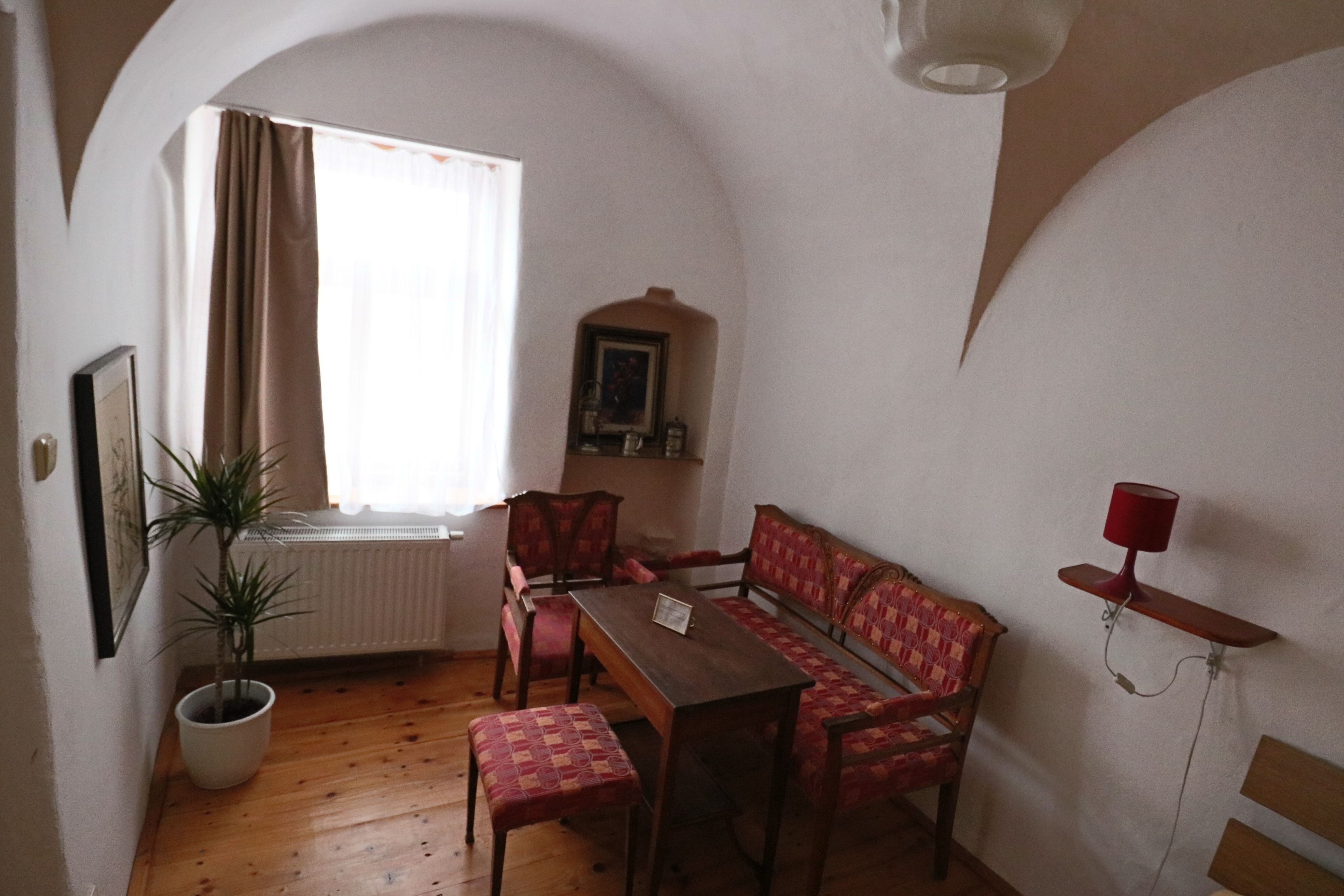 Una habitación acogedora con suelo de madera, una mesa de comedor con sillas, un banco, una planta en maceta y una lámpara roja sobre una estantería.