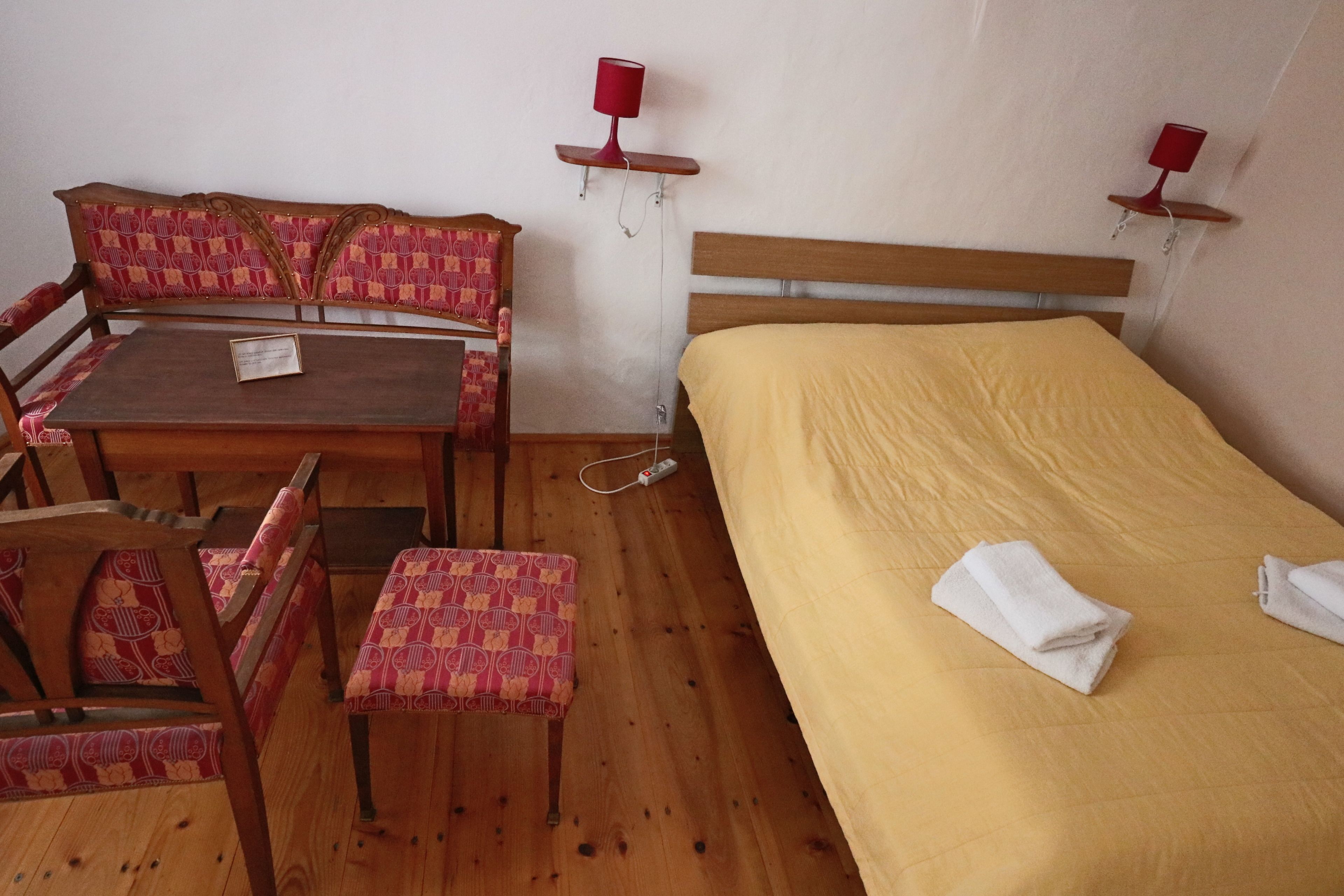Quarto com cama de madeira, mesa de madeira com cadeiras e duas luminárias fixadas na parede.