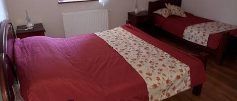 Un dormitorio con dos camas individuales, un radiador, una ventana con cortinas, una mesita de noche con lámpara y suelo de madera.
