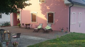 En lyserød bygning med en anlagt terrasse, to trædøre og et vindue med skodder. To loungesenge og et bord med stole står der.
