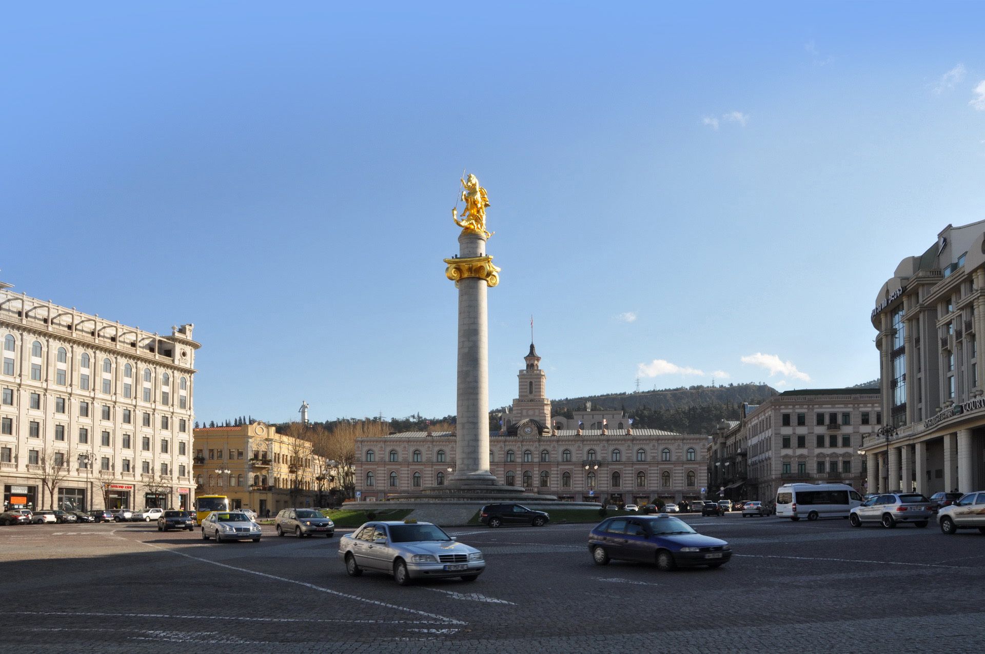 Een stadsplein met een hoog monument, omringd door historische gebouwen en een geplaveid straat.