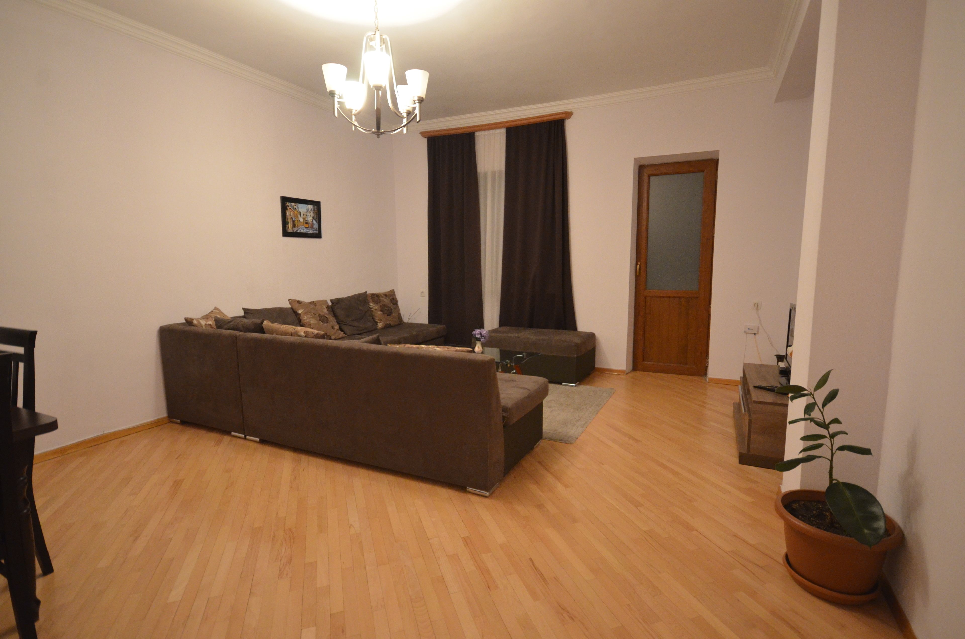 Un salon avec un canapé marron, un plancher en bois, un lustre, une porte et une plante en pot.