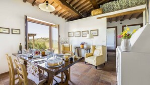 A dining area with a wooden table set for four, surrounded by wicker chairs. There is a view of the outdoors through a large window, and a staircase leading to an upper level. - Farmhouse in the Tuscan countryside (Semproniano)