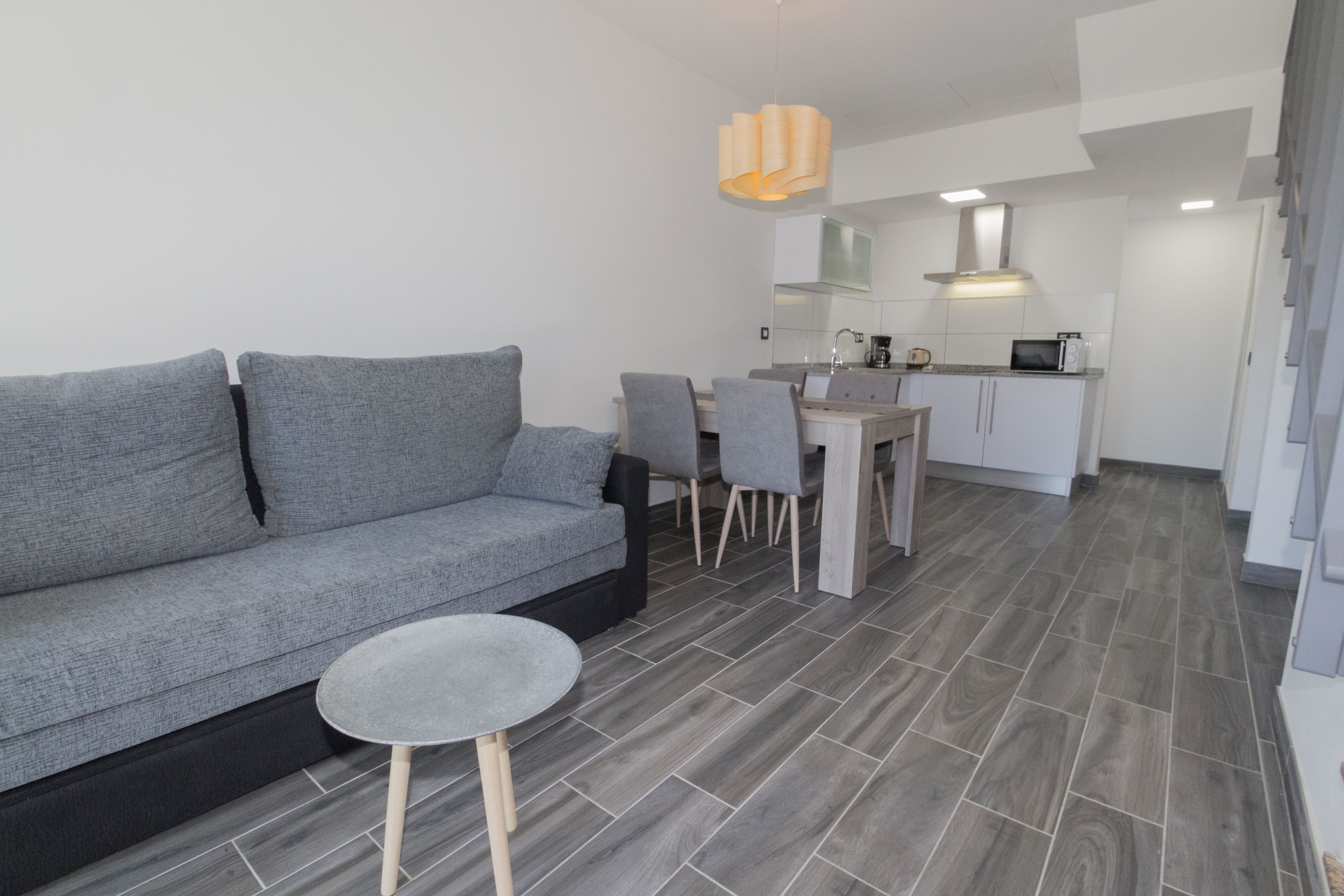 A modern living area with a grey sofa, a small round table, and a dining table with chairs. The room has a kitchen in the background with white cabinets and a microwave.