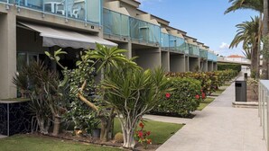 A row of hotel rooms with balconies, a paved walkway, and various plants and shrubs. - Bungalows Corinto II (San Bartolome de Tirajana)