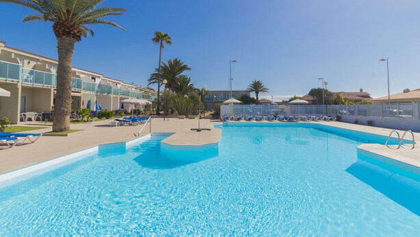 A clear blue swimming pool with lounge chairs and palm trees, surrounded by hotel buildings. - Bungalows Corinto II (San Bartolome de Tirajana)