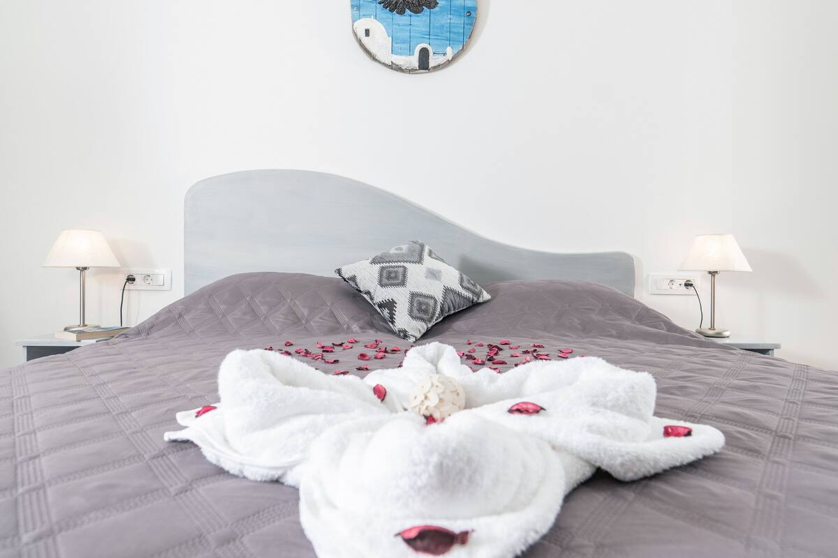 Ein ordentlich bezogenes Bett mit weißem Handtuch und Rosenblättern, einem gemusterten Kopfkissen und zwei Nachttischlampen.