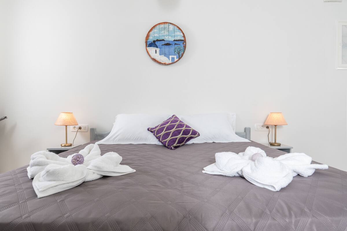 Ein ordentlich bezogenes Bett mit einem violetten Kissen, zwei Nachttischlampen und einem Bild darüber.