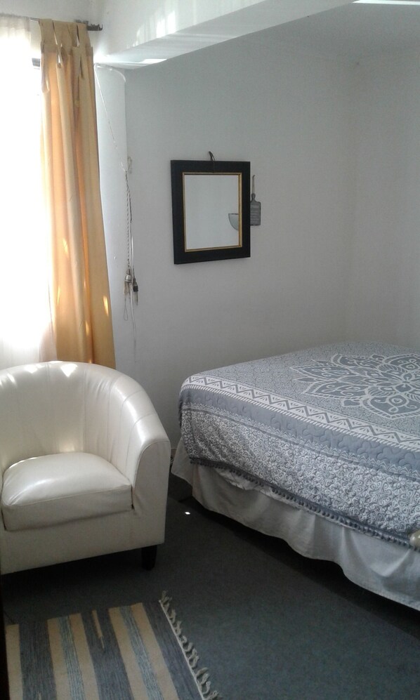 A small, simple room with a white armchair, a bed with patterned bedding, and a wall-mounted mirror.