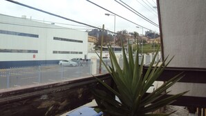 A view from a window with a plant in the foreground, a street with parked cars, and a building with a blue and white facade. - Habitaciones Florencia La Serena (La Serena)