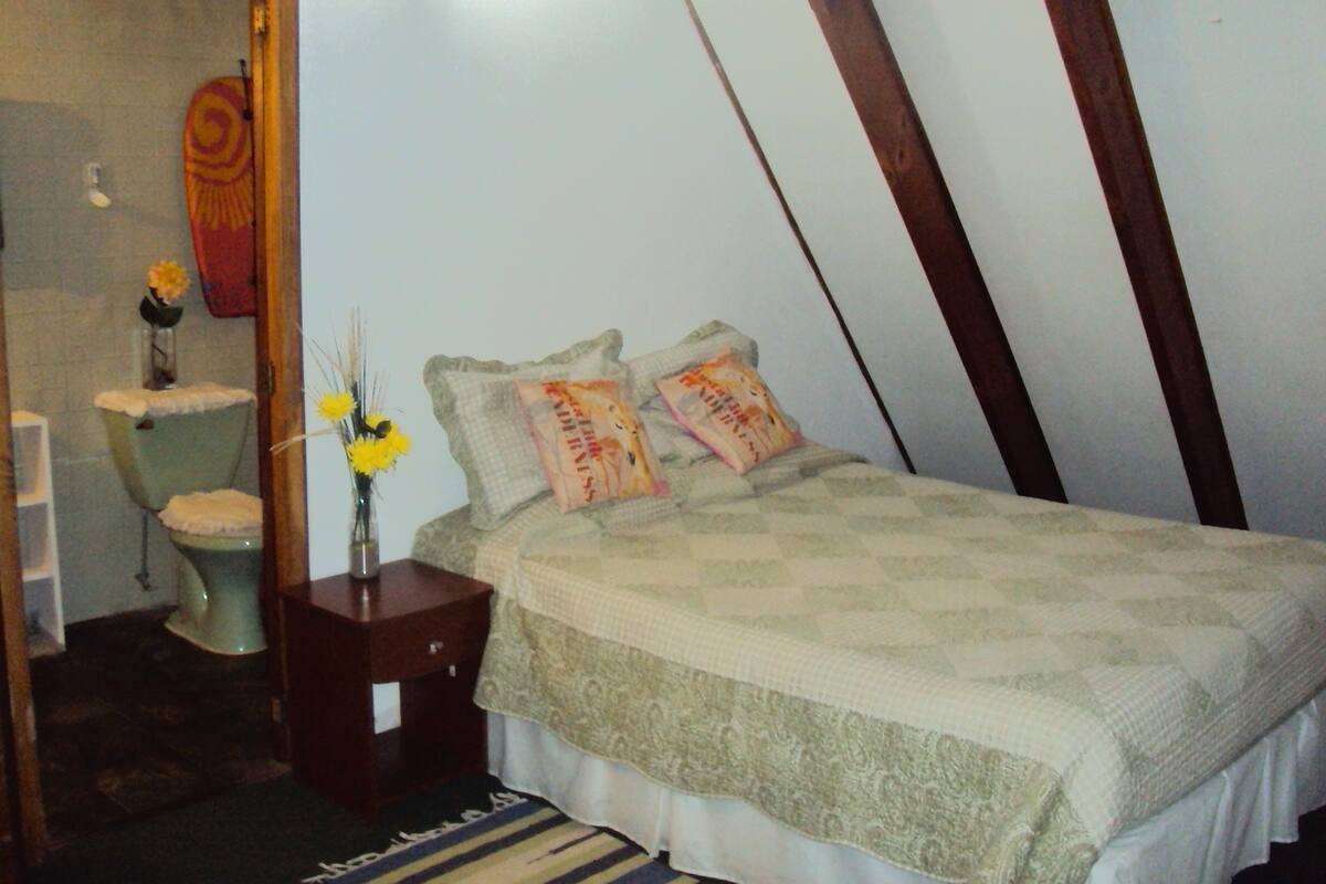 A bedroom with a bed, a bedside table, a nightstand, a small table with a vase of flowers, and a toilet.